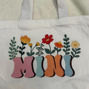 May include: A white canvas tote bag with two top handles featuring a colorful graphic print. The word MINI is written in large, blocky, pastel-colored letters, with various stylized flowers and green leaves growing out of the top of each letter.