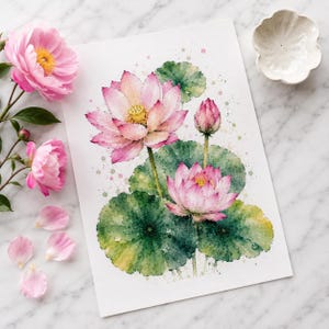 May include: A watercolor painting on white paper depicting two blooming pink lotus flowers, a closed bud, and large green lily pads with paint splatters. The artwork is surrounded by loose pink flower petals and a small white ceramic dish.