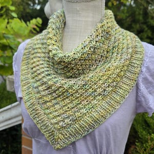 May include: A hand-knitted cowl scarf in shades of green and yellow, displayed on a mannequin. The scarf features a textured stitch pattern and a triangular shape, perfect for adding warmth and style.