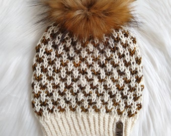 May include: A white and brown knitted beanie with a large brown faux fur pom pom. The beanie has a brown leather tag with the word "Mia" on it.