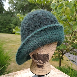 May include: A dark green, hand-knitted hat with a folded brim. The hat is displayed on a mannequin head with text from a "Harry Potter" book. The hat is made of a soft, textured yarn.