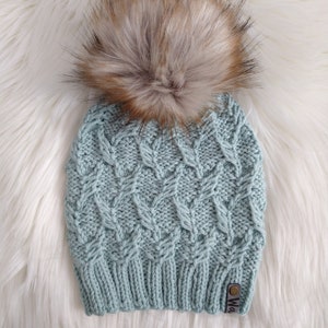 Może przedstawiać: Jasnoniebieska dzianinowa czapka z teksturowanym wzorem i dużym, puszystym, brązowo-beżowym pomponem ze sztucznego futra. Czapka ma małą metkę z nazwą marki. Czapka jest prezentowana na białej, puszystej powierzchni.