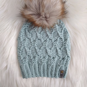 Może przedstawiać: Jasnoniebieska dzianinowa czapka z teksturowanym wzorem i dużym, puszystym, brązowo-beżowym pomponem ze sztucznego futra. Czapka ma małą, prostokątną metkę z nazwą marki. Czapka jest prezentowana na białej, puszystej powierzchni.