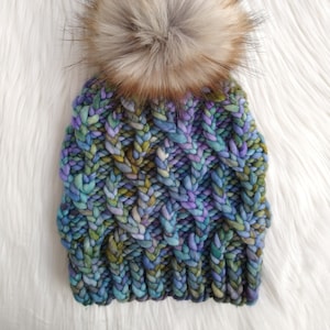 May include: A knitted beanie with a large, fluffy pom pom. The beanie is made in a multi-colored yarn with shades of blue, green, and purple.