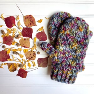 May include: A pair of hand-knitted mittens in a chunky, colorful yarn. The mittens feature a mix of blue, purple, yellow, and orange hues. Autumn leaves and acorns are arranged near the mittens on a white surface.
