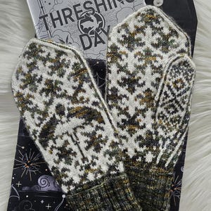 May include: A pair of knitted mittens featuring a white, green, and brown geometric pattern with ribbed cuffs, resting on a black fabric pouch with a bat print and a book titled Threshing Day with the subtitle Thirteen Unforgettable Stories.