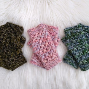 May include: Three pairs of knitted fingerless gloves in shades of green, pink, and brown. Each pair features a textured, woven pattern. The gloves are displayed on a white, fluffy surface, showcasing the handmade quality and color variations.