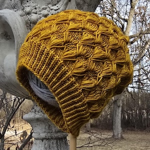 May include: A mustard yellow knitted beanie with a textured pattern. The beanie is made with a thick yarn and has a slouchy fit.