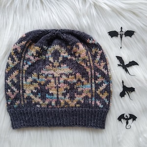 May include: A dark gray knitted hat with a colorful geometric pattern. The hat is on a white fluffy surface with black paper dragon cutouts.