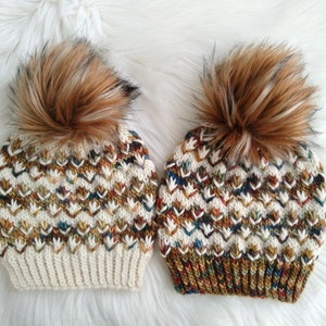 May include: Two knitted hats with brown faux fur pom poms. The hats have a white base with a chevron pattern in brown, gold, and blue.