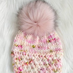 May include: A hand-knitted beanie in shades of pink, cream, and green, topped with a large, fluffy pink faux fur pom-pom. The hat features a chunky knit design with a folded brim. The background is a white, fluffy surface.