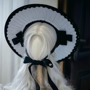 May include: A wide-brimmed hat featuring a pleated white fabric crown and brim, accented with a solid black outer edge and black ribbon bows on the sides. A black ribbon chin strap is tied in a bow beneath the base of the hat.