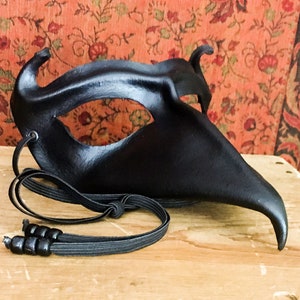 May include: A black leather masquerade mask with a long, pointed nose and a single eye hole. The mask has a black elastic strap.
