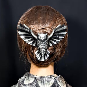May include: A silver owl hair clip, featuring detailed feathering and outstretched wings, set against brown hair. The clip is worn at the back of the head. The owl's wings are spread wide, creating a striking silhouette.