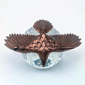 May include: A bronze-colored metal bird sculpture with outstretched wings, perched atop a clear glass orb. The bird's wings and tail feathers are detailed, and the sculpture has a metallic finish. The glass orb has bubbles inside.