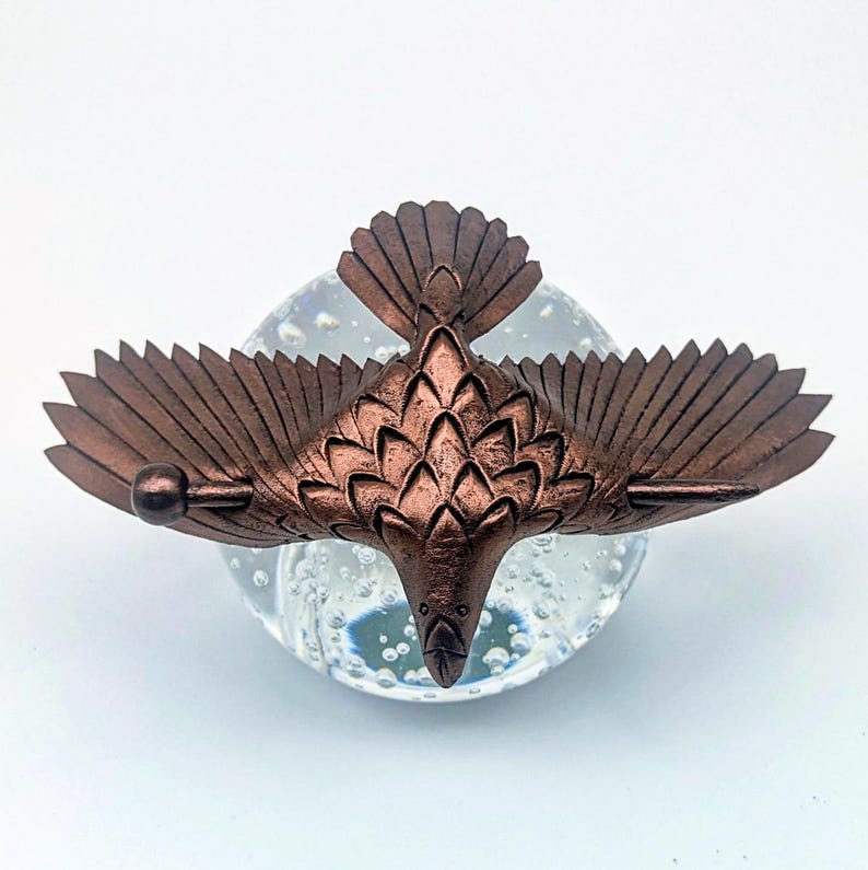 May include: A bronze-colored metal bird sculpture with outstretched wings, perched atop a clear glass orb. The bird's wings and tail feathers are detailed, and the sculpture has a metallic finish. The glass orb has bubbles inside.