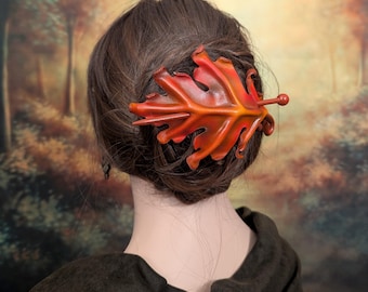 Könnte beinhalten: Ein Haaraccessoire in Form eines Eichenblatts mit einem Farbverlauf in Rot, Orange und Gelb. Das Blatt hat eine texturierte, geäderte Oberfläche und wird mit einem dünnen, passenden Stab befestigt, der durch das Haar gesteckt wird.