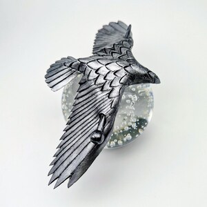 May include: A silver-colored, detailed sculpture of a bird in flight. The bird has outstretched wings and is perched on a clear glass orb with bubbles. The sculpture is set against a white background.