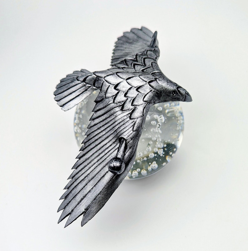 May include: A silver-colored, detailed sculpture of a bird in flight. The bird has outstretched wings and is perched on a clear glass orb with bubbles. The sculpture is set against a white background.