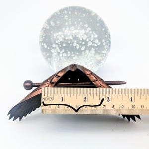 May include: A bronze-colored metal turtle-shaped object with a ruler showing a length of approximately 3 inches. The turtle has detailed wing and shell patterns. A clear glass sphere with bubbles is in the background.