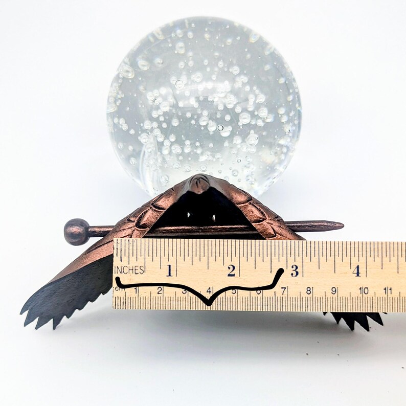 May include: A bronze-colored metal turtle-shaped object with a ruler showing a length of approximately 3 inches. The turtle has detailed wing and shell patterns. A clear glass sphere with bubbles is in the background.