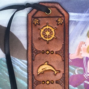 May include: A brown leather bookmark with a gold-colored design. The design features a ship's wheel, a dolphin, and a repeating pattern of circles and swirls.