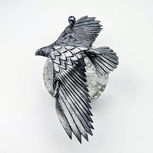 May include: A silver-colored eagle sculpture with outstretched wings, mounted on a clear glass orb. The eagle has detailed feather textures and is in mid-flight, creating a dynamic and artistic display. The sculpture is likely a decorative item.
