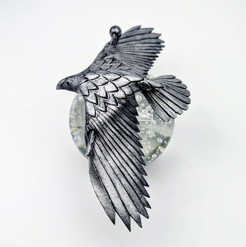 May include: A silver-colored eagle sculpture with outstretched wings, mounted on a clear glass orb. The eagle has detailed feather textures and is in mid-flight, creating a dynamic and artistic display. The sculpture is likely a decorative item.