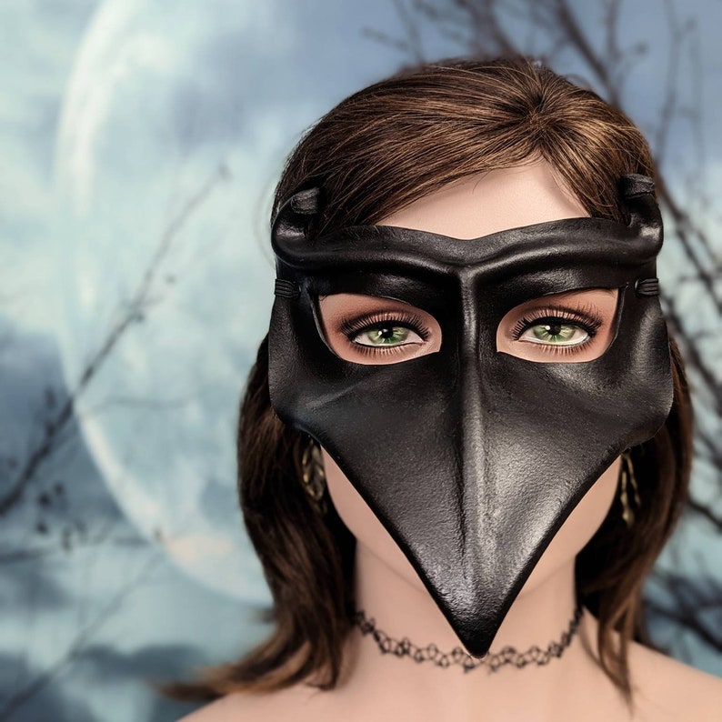 May include: A black leather masquerade mask with a long, pointed beak. The mask has green eye holes and is worn by a person with brown hair.