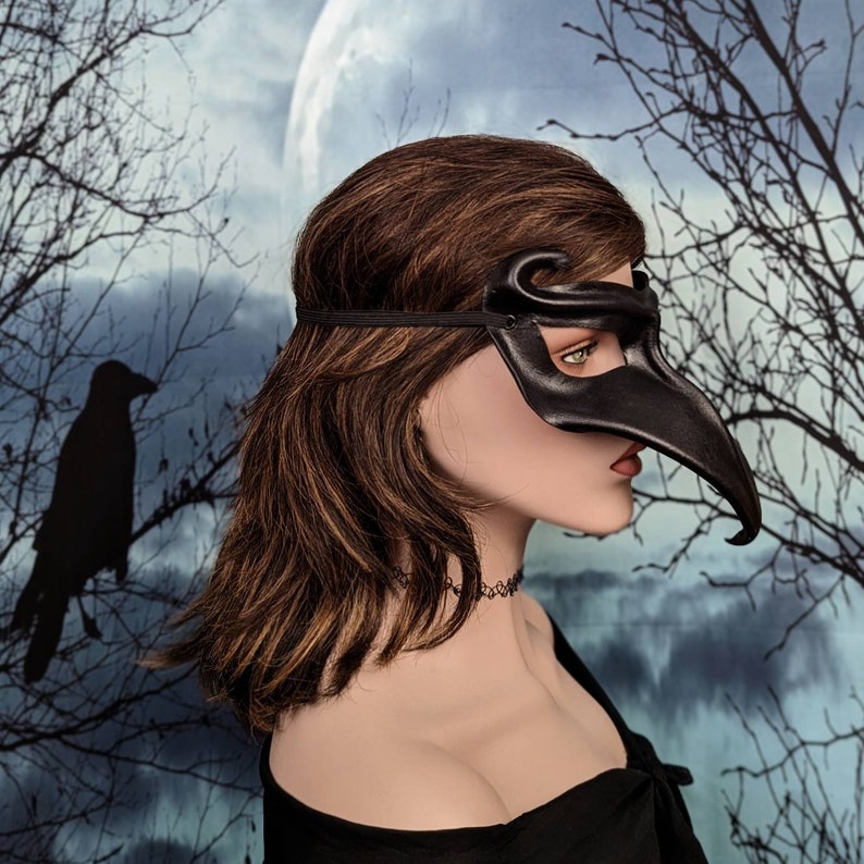 May include: A black leather masquerade mask with a long, curved beak. The mask is worn by a person with long brown hair. The background is a blurry image of a forest with a full moon.
