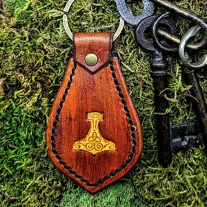 May include: A brown leather keychain with a gold Mjolnir hammer and a Celtic knot design. The keychain is attached to a silver key ring.