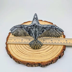 May include: A detailed, silver-colored raven sculpture with outstretched wings, approximately 7 inches wide. The bird is perched on a wooden slice, showcasing intricate feather detailing. The sculpture's wingspan is about 18 cm.