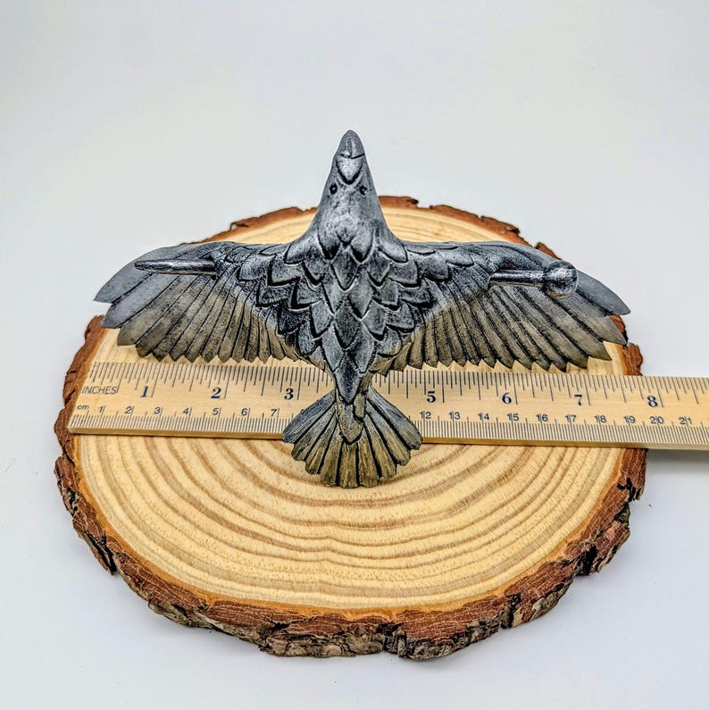 May include: A detailed, silver-colored raven sculpture with outstretched wings, approximately 7 inches wide. The bird is perched on a wooden slice, showcasing intricate feather detailing. The sculpture's wingspan is about 18 cm.