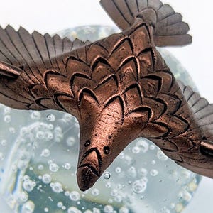 May include: A detailed bronze-colored bird sculpture in flight, with textured feathers and wings. The bird is set against a blurred background of bubbles, creating a sense of movement and depth. The sculpture's intricate details are highlighted by the light.
