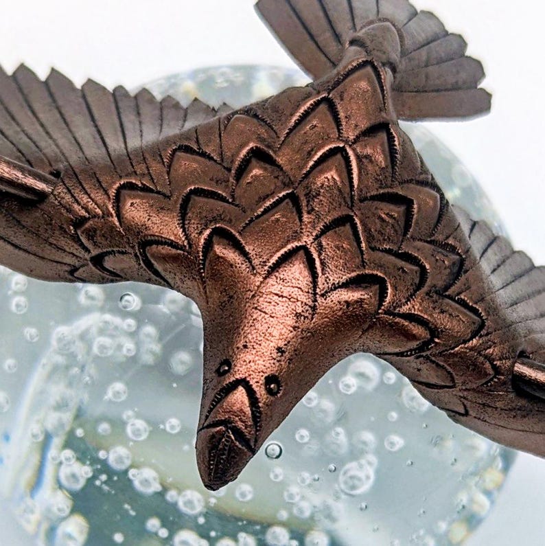 May include: A detailed bronze-colored bird sculpture in flight, with textured feathers and wings. The bird is set against a blurred background of bubbles, creating a sense of movement and depth. The sculpture's intricate details are highlighted by the light.