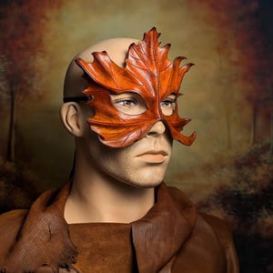 May include: A mask shaped like a large, textured maple leaf in shades of orange and brown. The mask covers the upper portion of the face, featuring cutouts for the eyes and a pointed, leaf-like structure that extends across the forehead.