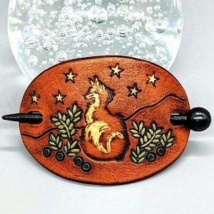 May include: A brown leather hair stick with a fox design and stars. The fox is outlined in gold and the stars are yellow. There are green leaves and blue berries around the fox.