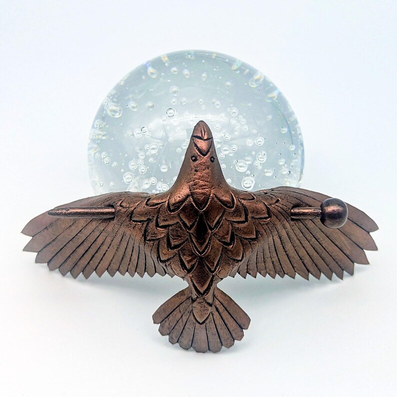 May include: A bronze-colored bird sculpture with outstretched wings, detailed feathers, and a round ball at the end of each wing. The bird is positioned in front of a clear glass sphere with bubbles.
