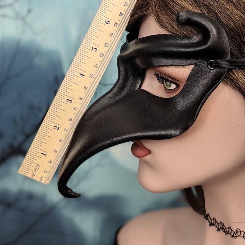 May include: A black leather masquerade mask with a long, curved beak. The mask is being held up to a ruler, showing that the beak is approximately 15 cm long.