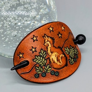 K&ouml;nnte beinhalten: Ein brauner Haarstecker aus Leder mit einem Fuchsdesign und einem Sternenmuster. Der Fuchs ist in Gold umrandet und die Sterne sind goldfarben mit einem wei&szlig;en Zentrum. Der Haarstecker hat einen schwarzen Holzstift.