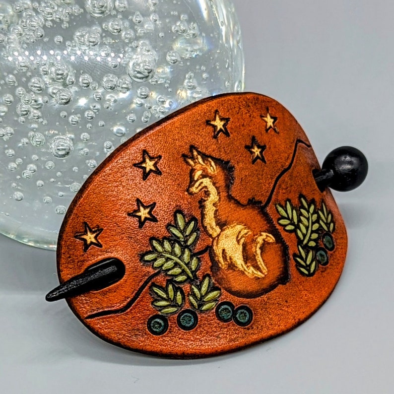 K&ouml;nnte beinhalten: Ein brauner Haarstecker aus Leder mit einem Fuchsdesign und einem Sternenmuster. Der Fuchs ist in Gold umrandet und die Sterne sind goldfarben mit einem wei&szlig;en Zentrum. Der Haarstecker hat einen schwarzen Holzstift.