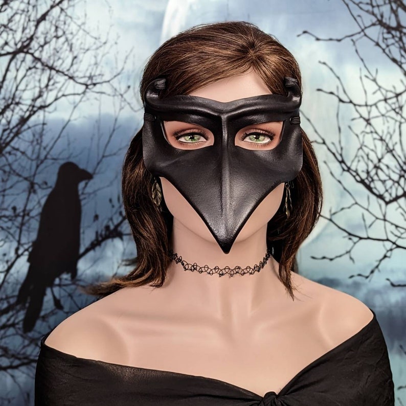 May include: A black leather masquerade mask with a long, pointed beak. The mask has eye holes and is decorated with a small, dangling ornament on each side. The mask is worn by a person with brown hair.