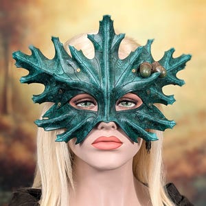 May include: A dark teal mask shaped like a large maple leaf with prominent veins and jagged edges. The mask features two small, realistic brown acorns attached to the upper right side and small metallic rivets securing the leaf-like structure.