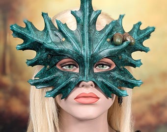 Könnte beinhalten: Eine dunkel-türkisfarbene Maske in Form eines großen Ahornblatts mit ausgeprägten Blattadern und gezackten Rändern. Die Maske ist mit zwei kleinen, realistischen braunen Eicheln auf der rechten Seite und kleinen Metallnieten versehen.
