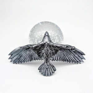 May include: A silver-colored raven sculpture with outstretched wings, detailed feathers, and a textured surface. The raven is positioned in front of a clear, spherical object with internal bubbles, set against a white background.