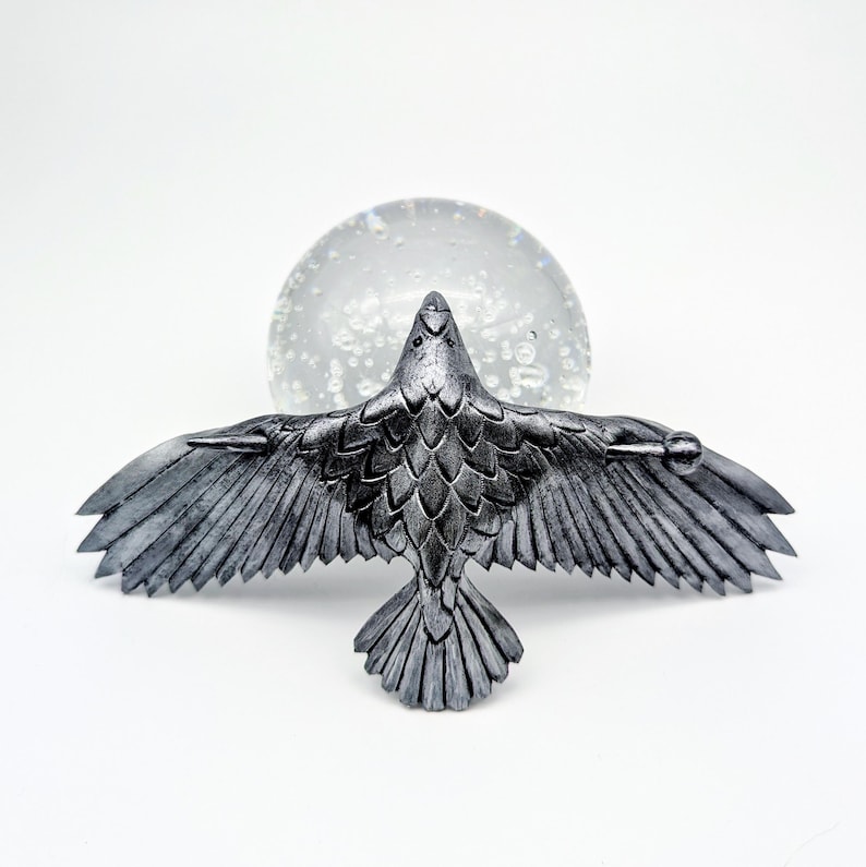 May include: A silver-colored raven sculpture with outstretched wings, detailed feathers, and a textured surface. The raven is positioned in front of a clear, spherical object with internal bubbles, set against a white background.
