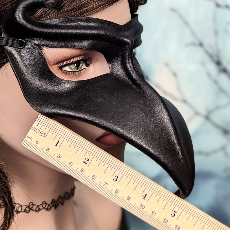 May include: A black leather masquerade mask with a long, pointed beak. The mask is being held up to a ruler, showing that it is approximately 12.7 cm long.