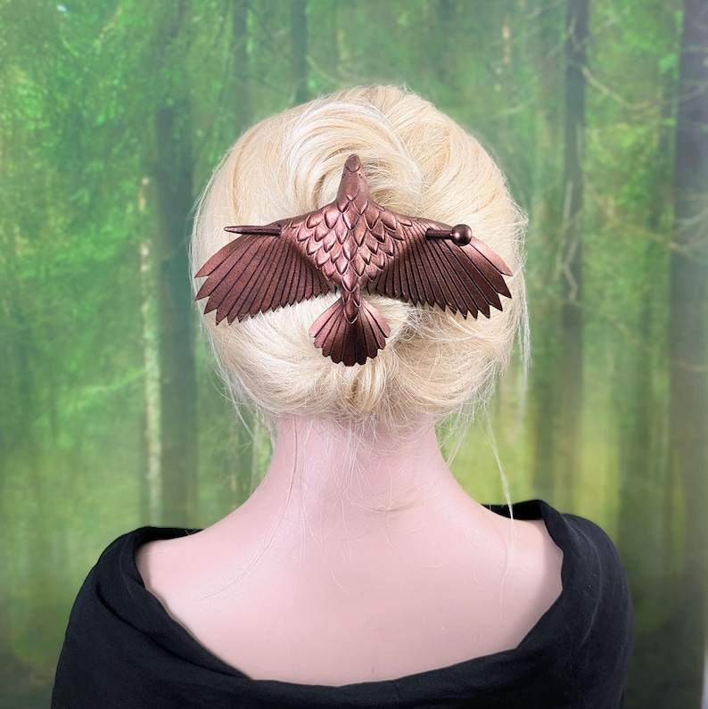 May include: A bronze-colored hair clip in the shape of a bird with outstretched wings. The hair accessory is attached to a bun of blonde hair. The bird's wings are detailed with individual feathers.