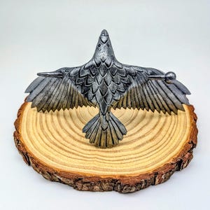 May include: A detailed, silver-colored raven sculpture with outstretched wings, perched on a natural wood slice. The raven's feathers are intricately carved, and the wood slice has a visible grain and bark edge. The sculpture is a decorative item.