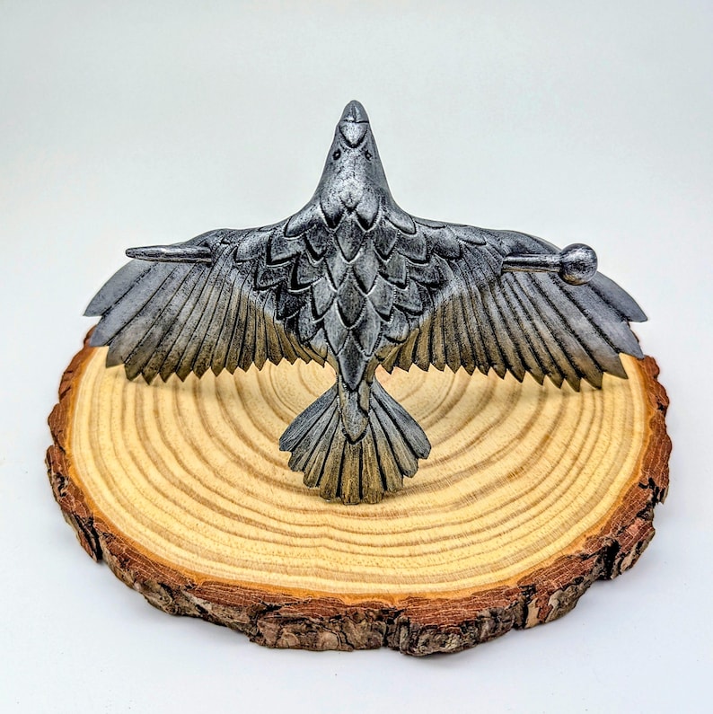 May include: A detailed, silver-colored raven sculpture with outstretched wings, perched on a natural wood slice. The raven's feathers are intricately carved, and the wood slice has a visible grain and bark edge. The sculpture is a decorative item.